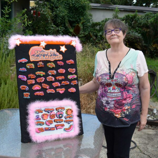 Young woman in a denim shirt placing a handmade Showgirl magnet onto a retro turquoise fridge. She is also wearing another Showgirl pin on her shirt, surrounded by other colorful polymer clay designs from Jeanette Meyer’s Showgirl Collection.
