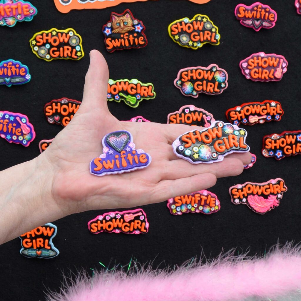 Close-up of a hand holding two handmade polymer clay pins from Jeanette Meyer’s Showgirl Collection: one reading Swiftie with a heart design and the other Showgirl with colorful accents, surrounded by additional pins displayed on a black background with pink feather trim.